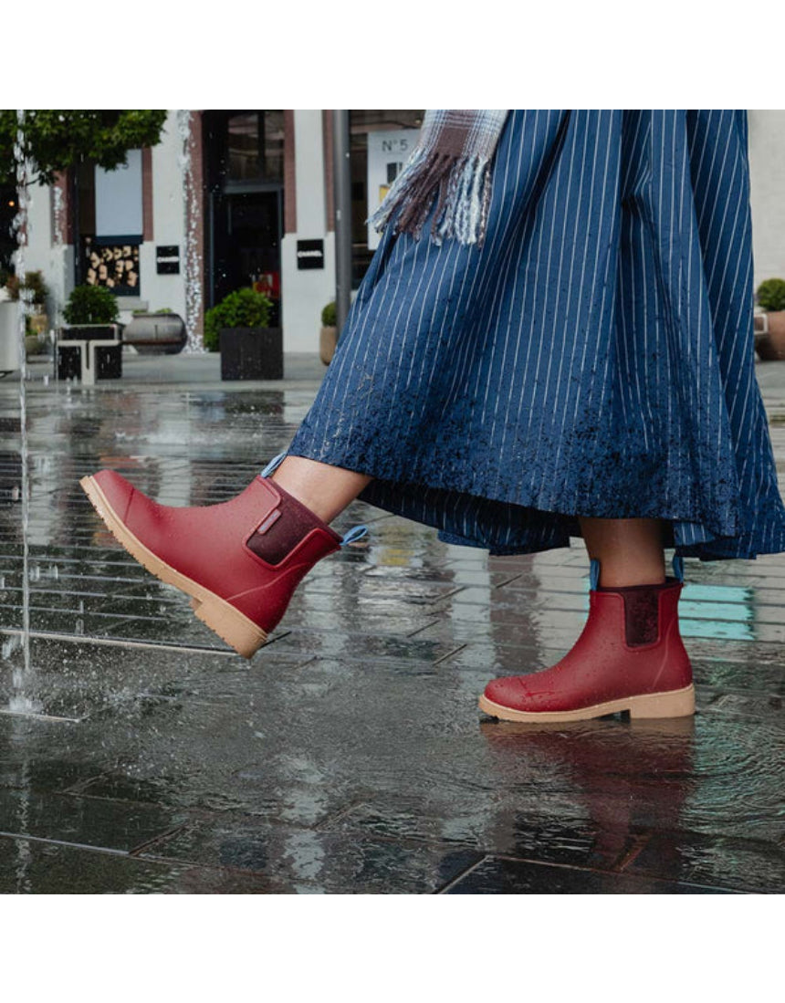 Brick red Merry People Bobbi gumboots shown in a lifestyle setting with a person walking outdoors in wet weather.