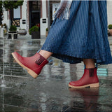 Brick red Merry People Bobbi gumboots shown in a lifestyle setting with a person walking outdoors in wet weather.