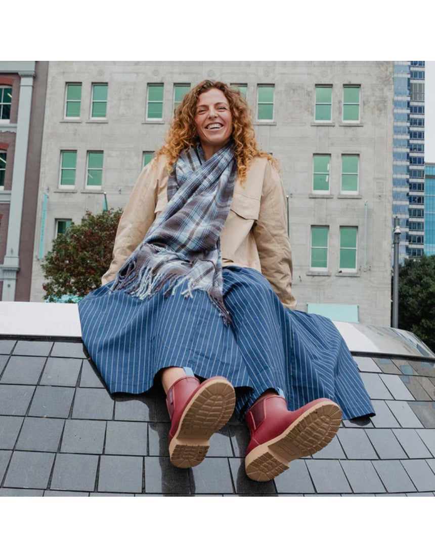 Merry People Bobbi gumboots in brick red, outdoor lifestyle image showing waterproof rubber boots on a person walking in a muddy area.